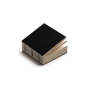 May include: A stack of small, square notebooks with black covers and cream-colored pages. The pages are bound together with visible stitching along the spine. The notebooks are stacked neatly, with a few pages of one notebook slightly fanned open.