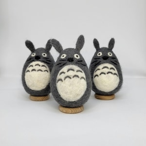May include: Three gray felt Totoro figurines with white faces and black eyes. Each figurine is sitting on a wooden base.