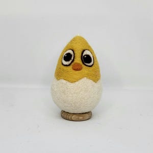 Op de afbeelding: Een geel vilt kuiken met zwarte ogen en een kleine oranje snavel kijkt uit een witte vilt eierschaal. Het kuiken staat op een kleine houten basis.