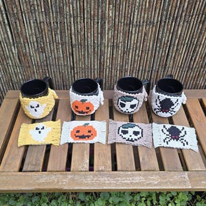 May include: Four black ceramic mugs each featuring a unique macrame cozy, accompanied by four matching rectangular macrame coasters. The designs include a yellow ghost, an orange pumpkin, a beige skull, and a black spider on a white background.