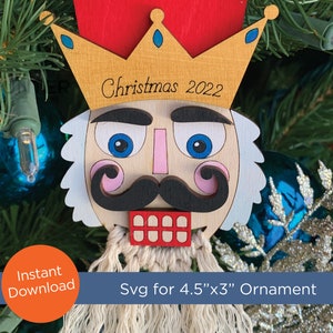 Puede incluir: Adorno navideño de madera con forma de cascanueces, con una corona dorada y el texto "Christmas 2022". El adorno tiene una parte superior roja, una cara de madera clara y un bigote negro. El adorno mide aproximadamente 11,4 cm por 7,6 cm.