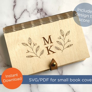 Puede incluir: Una cubierta de libro de madera con un diseño cortado con láser que presenta las iniciales "M K" y una rama frondosa. La cubierta incluye una correa de cuero con una hebilla de latón. El texto "Incluye diseño para marcar" es visible en la esquina superior derecha. El texto "Descarga instantánea" es visible en la esquina inferior izquierda. El texto "SVG/PDF para cubierta de libro pequeña" es visible en el centro inferior.