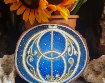 May include: A hand-painted wooden slice with a celestial design. The artwork features a blue background with white stars and a gold and white geometric pattern. The wood has a natural bark edge.