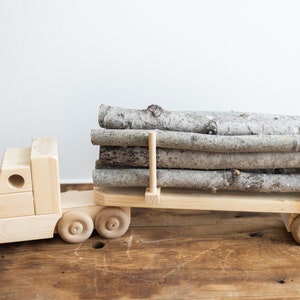 May include: A wooden toy truck with a flatbed trailer carrying a load of logs. The truck is made of natural wood and has a simple design. The logs are stacked neatly on the trailer and are secured with wooden dowels.