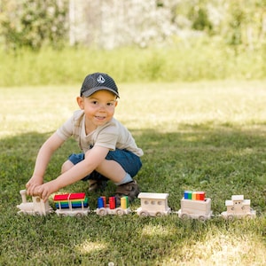 May include: A young boy wearing a black baseball cap plays with a wooden toy train set on a grassy lawn. The train has five carriages, including an engine, a flatbed carriage, a boxcar, and two passenger carriages. The train is painted in natural wood tones and features colourful accents.