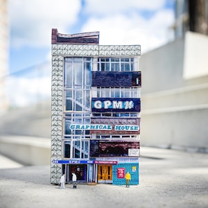 May include: A miniature model of a building with a blue and white sign that reads "GPM II GRAPHICAL HOUSE". The building is made of paper and has a brick facade. There are two small figures standing in front of the building.