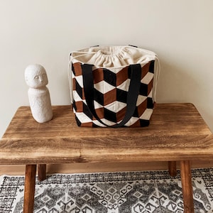 Op de afbeelding: Een tote bag met een geometrisch patroon van zwarte, bruine en witte ruitvormen. De tas heeft zwarte handvatten en een beige trekkoordsluiting. De tas staat op een houten tafel, met links een klein wit beeld.
