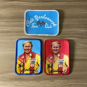 May include: Three embroidered patches. The top patch is light blue with white stitching and the text "Cale Yarborough Fan Club" with a checkered flag graphic. The bottom two patches are rectangular and feature a photo of Cale Yarborough in a racing suit with his signature below. The left patch is blue with white stitching and the right patch is red with white stitching.