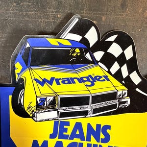K&ouml;nnte beinhalten: Ein gestanztes Schild mit einem gelben und blauen Rennwagen, dem Schriftzug Wrangler auf der Motorhaube und den Worten JEANS MACHINE in gro&szlig;en blauen Buchstaben darunter. Hinter der Autoillustration befindet sich eine schwarz-wei&szlig;e Zielflagge.