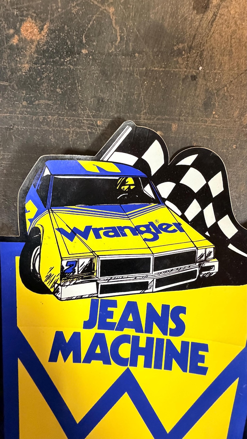 K&ouml;nnte beinhalten: Ein gestanztes Schild mit einem gelben und blauen Rennwagen, dem Schriftzug Wrangler auf der Motorhaube und den Worten JEANS MACHINE in gro&szlig;en blauen Buchstaben darunter. Hinter der Autoillustration befindet sich eine schwarz-wei&szlig;e Zielflagge.
