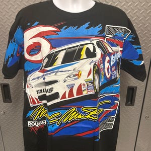 Puede incluir: Camiseta negra con un diseño de coche de carreras. La camiseta presenta el número 6 y el nombre Mark Martin. El coche es blanco, rojo y azul, con logotipos de patrocinadores. Tiene cuello redondo y mangas cortas.
