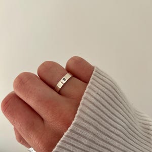 May include: A silver ring with the letter "B" engraved on it. The ring is worn on the index finger of a hand.
