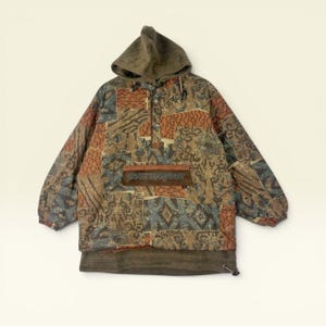 Könnte beinhalten: Ein langärmeliger Kapuzenpullover mit Patchwork-Muster in Braun-, Orange- und Blautönen. Das Kleidungsstück verfügt über eine Kapuze, eine Kängurutasche vorne mit dem auf einem braunen Aufnäher gestickten Text "courmayeur" und einen Kordelzug am Saum.