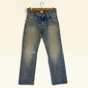 May include: A pair of blue denim jeans featuring a faded wash, a button fly, and a distressed rip on the right knee. The jeans have a classic five-pocket design with copper-colored rivets, belt loops, and a straight-leg cut hanging on a wooden hanger.