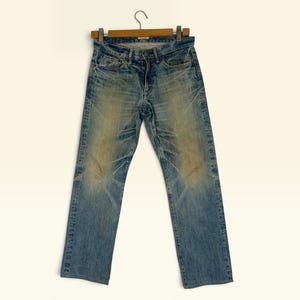 May include: Blue denim jeans featuring a faded wash with whiskering patterns at the hips and thighs. The jeans have a classic five-pocket design, a button fly closure, and belt loops, displayed hanging on a wooden hanger against a plain background.