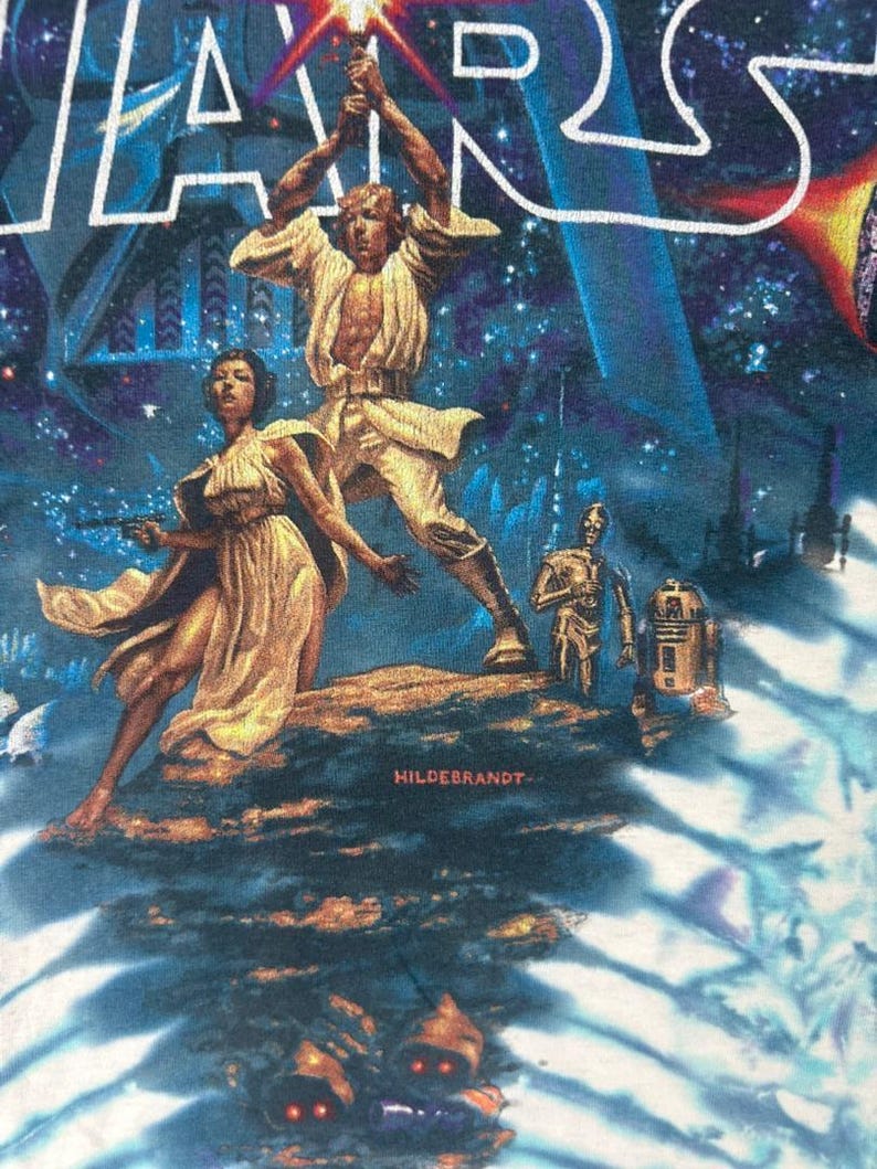 Puede incluir: Una camiseta gr&aacute;fica de Star Wars con una vibrante ilustraci&oacute;n de Luke Skywalker empu&ntilde;ando un sable de luz, con la Princesa Leia, C-3PO y R2-D2 de fondo. La palabra "STAR WARS" est&aacute; en la parte superior en azul y blanco.