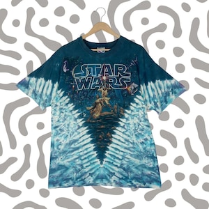 Puede incluir: Una camiseta de Star Wars te&ntilde;ida con un patr&oacute;n azul y blanco. La camiseta presenta el logotipo de Star Wars y un gr&aacute;fico de personajes de la pel&iacute;cula. La camiseta est&aacute; colgada en una percha de madera.