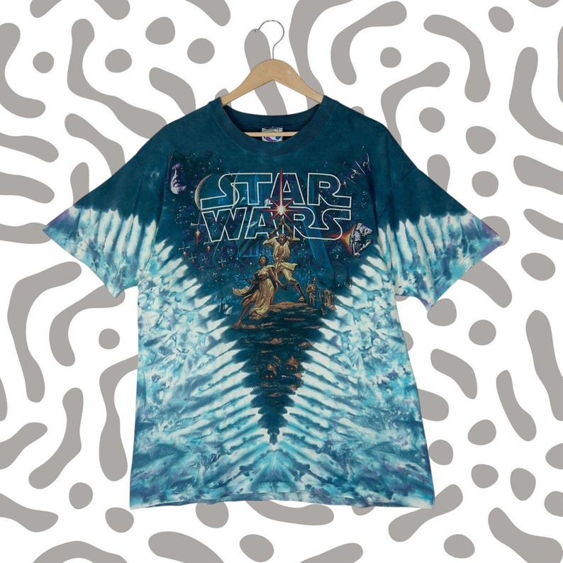 Puede incluir: Una camiseta de Star Wars te&ntilde;ida con un patr&oacute;n azul y blanco. La camiseta presenta el logotipo de Star Wars y un gr&aacute;fico de personajes de la pel&iacute;cula. La camiseta est&aacute; colgada en una percha de madera.