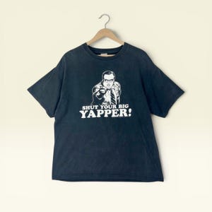 Op de afbeelding: Een zwart t-shirt met korte mouwen en een witte grafische print van een persoon met een bril en een pak die naar voren wijst. Onder de afbeelding staat de tekst SHUT YOUR BIG YAPPER! in dikke, witte hoofdletters. Het shirt hangt aan een houten hanger.
