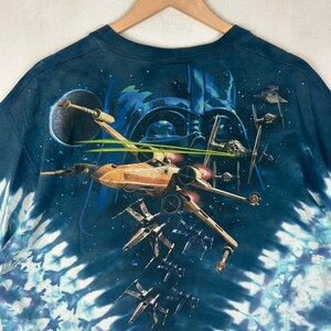 Puede incluir: Camiseta azul marino con dise&ntilde;o tie-dye y motivo de Star Wars. El gr&aacute;fico incluye cazas X-wing, cazas TIE y la silueta de Darth Vader sobre un fondo estrellado. La camiseta est&aacute; colgada en una percha de madera.