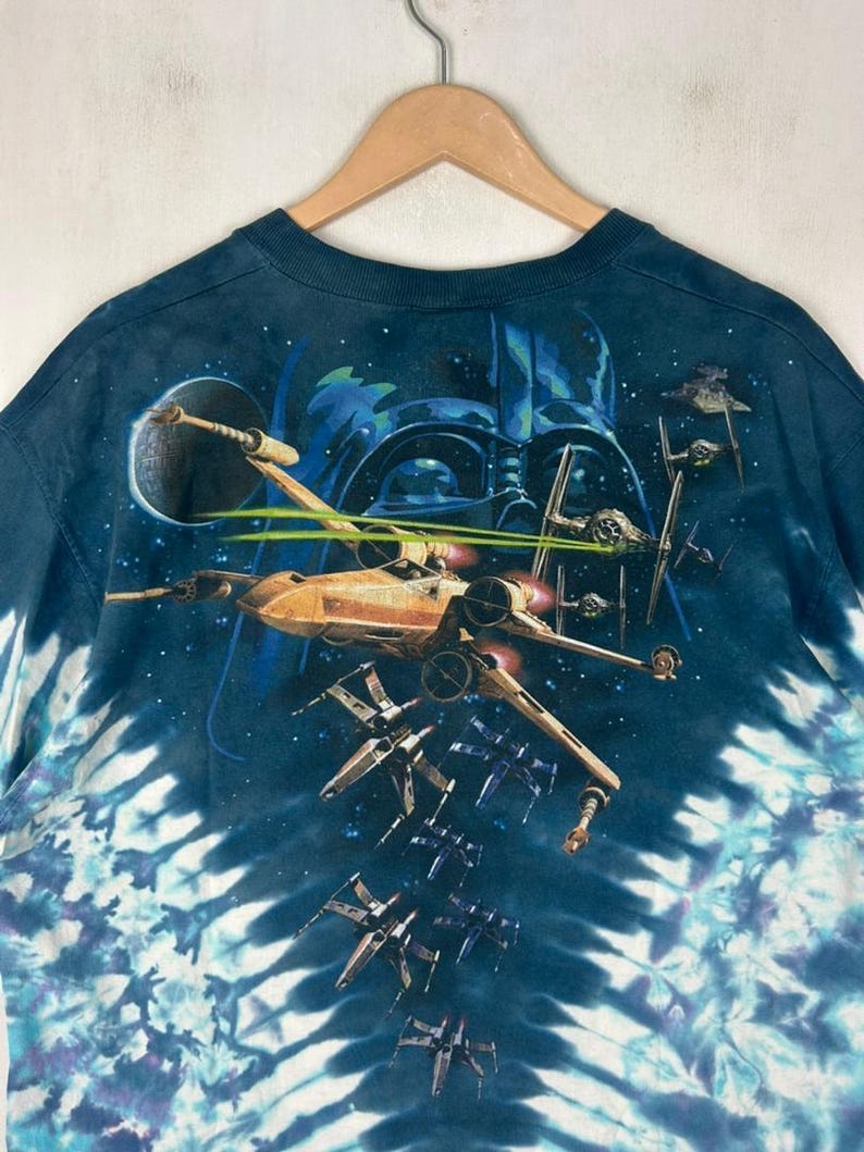 Puede incluir: Camiseta azul marino con dise&ntilde;o tie-dye y motivo de Star Wars. El gr&aacute;fico incluye cazas X-wing, cazas TIE y la silueta de Darth Vader sobre un fondo estrellado. La camiseta est&aacute; colgada en una percha de madera.