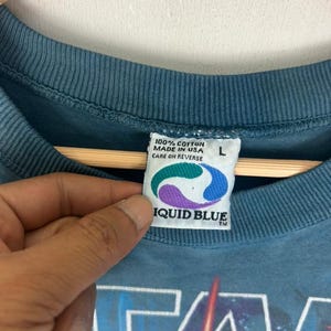 Puede incluir: Camiseta de color turquesa con un dise&ntilde;o gr&aacute;fico de Star Wars. La etiqueta indica 100% algod&oacute;n, fabricado en EE. UU. y Liquid Blue. Talla L. Instrucciones de cuidado al reverso.