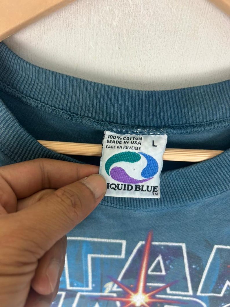 Puede incluir: Camiseta de color turquesa con un dise&ntilde;o gr&aacute;fico de Star Wars. La etiqueta indica 100% algod&oacute;n, fabricado en EE. UU. y Liquid Blue. Talla L. Instrucciones de cuidado al reverso.