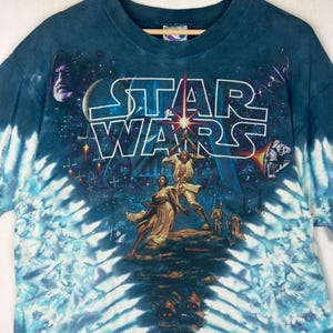 Puede incluir: Una camiseta de Star Wars azul te&ntilde;ida con un dise&ntilde;o de personajes y el logotipo de Star Wars. La camiseta tiene cuello redondo y mangas cortas. El dise&ntilde;o incluye im&aacute;genes de Darth Vader, Luke Skywalker y la Princesa Leia.
