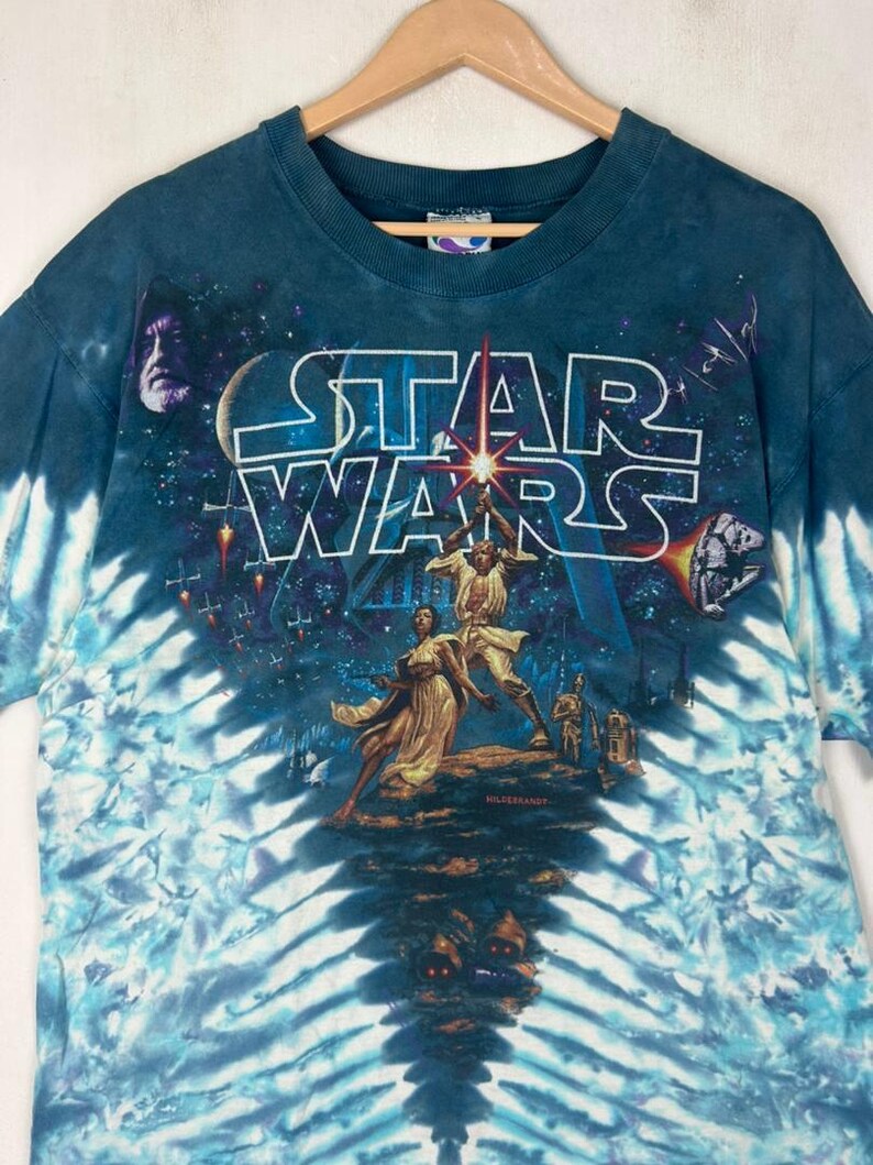 Puede incluir: Una camiseta de Star Wars azul te&ntilde;ida con un dise&ntilde;o de personajes y el logotipo de Star Wars. La camiseta tiene cuello redondo y mangas cortas. El dise&ntilde;o incluye im&aacute;genes de Darth Vader, Luke Skywalker y la Princesa Leia.