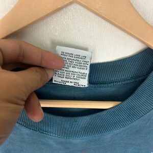 Puede incluir: Primer plano de una camiseta de color verde azulado con un gr&aacute;fico de Star Wars. La etiqueta de la prenda muestra las instrucciones de cuidado y "MADE IN USA". La camiseta est&aacute; colgada en una percha de madera.