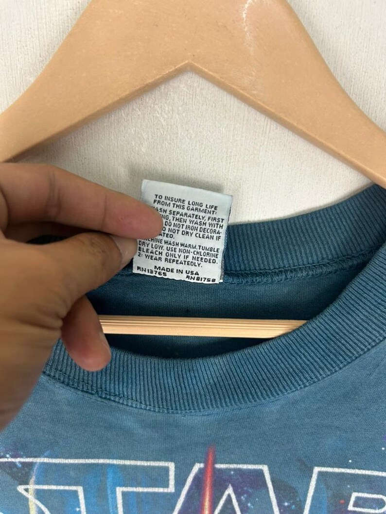 Puede incluir: Primer plano de una camiseta de color verde azulado con un gr&aacute;fico de Star Wars. La etiqueta de la prenda muestra las instrucciones de cuidado y "MADE IN USA". La camiseta est&aacute; colgada en una percha de madera.