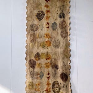 May include: A long, beige fabric table runner with a scalloped edge, decorated with leaf and floral imprints in shades of brown, green, and orange. The runner is hanging on a white wall, suspended from a clothes hanger.