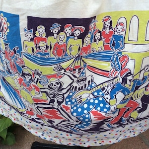 Op de afbeelding: Een witte stof met een kleurrijke print van een carnavals scène. De print toont een skelet, een clown, een muzikant en een groep mensen in kostuums. De stof heeft een witte achtergrond met gele, blauwe, rode en zwarte accenten.