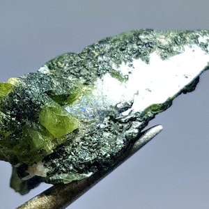 Puede incluir: Un grupo de cristales verdes incrustados en una matriz gris oscura. Los cristales tienen un brillo vítreo y son translúcidos. La matriz es rugosa y texturizada.