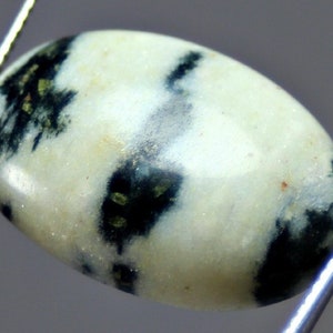 May include: A white oval cabochon gemstone with black and green inclusions. The stone is smooth and polished.