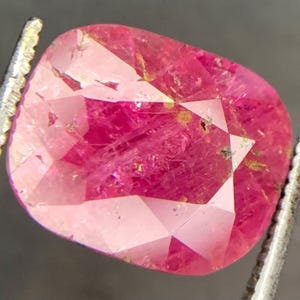May include: A faceted, cushion-cut ruby gemstone. The gem is a vibrant pinkish-red color, with visible inclusions and imperfections. The stone is held by a pair of metal tweezers, showcasing its shape and facets. The ruby's surface reflects light, highlighting its cut.