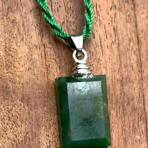 Könnte beinhalten: Ein rechteckiger, dunkelgrüner Jade-Anhänger mit silberfarbener Öse hängt an einer gedrehten grünen Kordel. Der Jade hat eine polierte Oberfläche. Der Anhänger ist ca. 2,5 cm lang, vor einem Holzgrund.