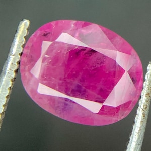 May include: A faceted, oval ruby gemstone. The gem is a vibrant pinkish-red colour, with internal inclusions and a polished surface. The ruby is held by silver tweezers against a dark background. The gemstone is the focal point of the image.