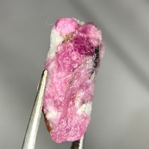 May include: A close-up of a rough, uncut ruby gemstone. The ruby is held by tweezers, showcasing its vibrant pink and white coloration with dark inclusions. The stone has a textured surface.