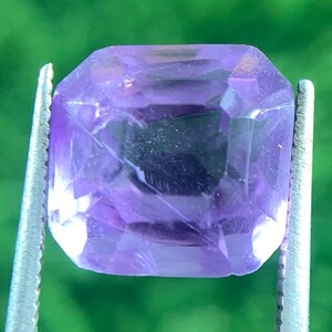 Könnte beinhalten: Ein facettierter, rechteckig geschliffener Amethyst-Edelstein, der von einer silbernen Pinzette gehalten wird. Der Edelstein hat eine leuchtende lila Farbe mit inneren Einschlüssen. Der Hintergrund ist ein verschwommenes Grün.