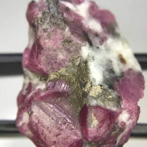 May include: A pink ruby crystal with white and gray inclusions. The crystal is rough and has a natural, unpolished look.