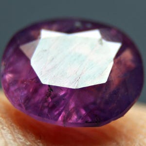 Può includere: Primo piano di una gemma sfaccettata di forma ovale e di colore viola intenso. La gemma presenta un'ampia zona centrale bianca multifattoriale, circondata da un corpo traslucido rosso-viola. La pietra è posta su uno sfondo neutro.