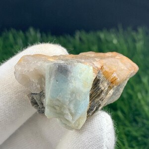 May include: A raw, natural gemstone with a light blue and beige color palette. The stone has a rough, textured surface and is held in a white gloved hand. The background is a mix of green and dark colors.