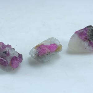 May include: Three rough ruby specimens with a mix of pink and white coloration. The rubies have a crystalline structure and are partially embedded in a white matrix. The stones vary in size and shape, with some showing dark gray inclusions.