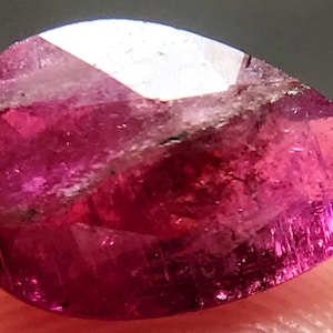 May include: A pear-shaped, faceted gemstone with a deep pink hue and visible internal inclusions. The gemstone has a high polish and a translucent appearance.