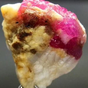 May include: A rough, pink ruby crystal embedded in a white and yellow matrix. The ruby has a deep, vibrant pink color and is surrounded by a white, crystalline matrix. The matrix is also speckled with yellow and brown areas.