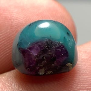 Pode incluir: Uma pedra preciosa tipo cabochão em forma de cúpula, apresentando uma seção superior azul turquesa vibrante e um centro roxo profundo e texturizado. A pedra tem um acabamento polido e brilhante com inclusões naturais visíveis.