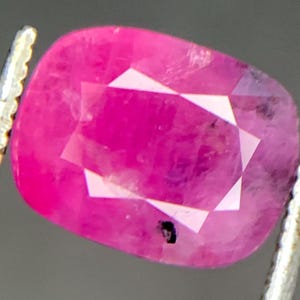 Op de afbeelding: Een gefacetteerde, ovaalvormige robijn edelsteen, vastgehouden door een zilveren pincet. De robijn heeft een levendige roze-rode kleur met een geometrisch patroon van facetten. De edelsteen heeft een paar donkere insluitsels.