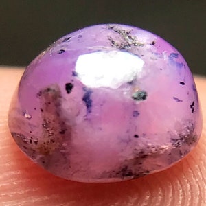 May include: A round, purple gemstone with a cabochon cut. The stone has a light purple color with dark purple and gray inclusions.