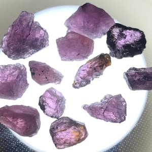 Pode incluir: Uma coleção de onze pedras preciosas brutas e não polidas de várias formas e tamanhos. As pedras exibem qualidades translúcidas a opacas com bordas naturais irregulares e tons violetas profundos sobre um fundo branco circular.